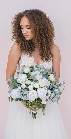 a woman in a wedding dress holding a bouquet of white flowers and greenery with her eyes closed