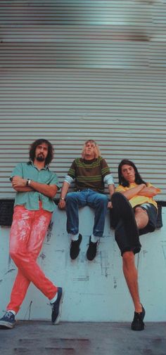 three men are sitting on a wall with their arms crossed and one man is leaning against the wall