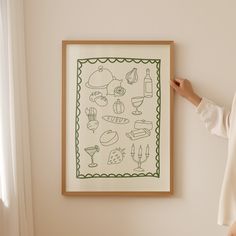 a woman standing in front of a white wall holding up a framed art print with food items on it