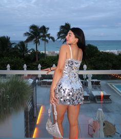 a woman in a silver dress looking out at the ocean and palm trees with her hand on her hip