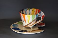 a cup and saucer on a plate with paintbrushes next to it in front of a gray background