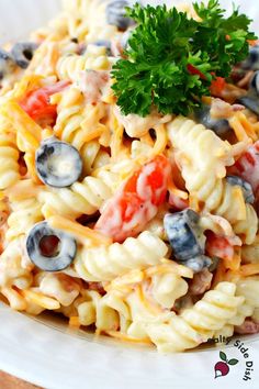 a pasta salad with tomatoes, olives and blueberries in a white bowl on a wooden table