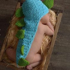 a baby sleeping in a box wearing a blue and green crochet dragon hat