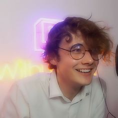 a young man wearing glasses and smiling in front of a wall mounted clock with neon lights behind him