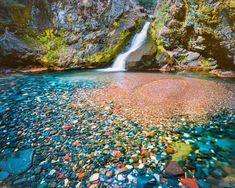 a small waterfall surrounded by lots of rocks and pebbles in the water with a rainbow colored stream running through it