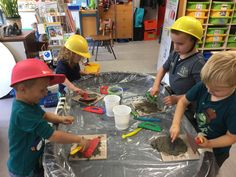 three children in hard hats are playing with sand and shovels on a plastic table