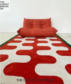 a red and white area rug with an orange pillow sitting on top of it in front of a white wall