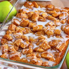 a glass baking dish filled with cinnamon rolls and apple slices next to green apples on a wooden table