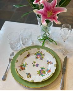 a table set with plates, silverware and pink lilies in a glass vase