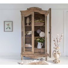 an old wooden cabinet with glass doors and shelves in the corner, next to a vase filled with flowers