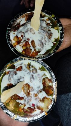 two metal pans filled with food covered in white sauce and toppings, being held by someone's hand