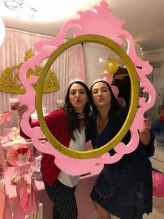 two women standing in front of a mirror with pink and gold decorations on the wall