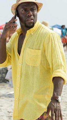 a man in yellow shirt and hat on cellphone at beach with umbrellas behind him