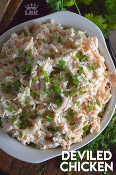 a white bowl filled with chicken salad on top of a wooden table next to parsley