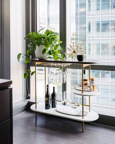 a bar cart with bottles and glasses on it in front of a window next to a potted plant