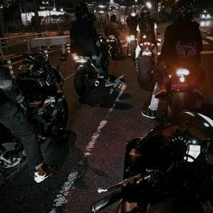 a group of people riding motorcycles down a street at night with their headlights on and one person standing next to them