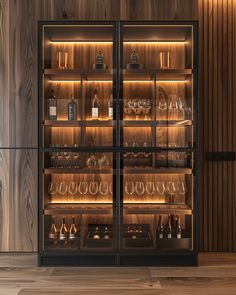 an illuminated display case with wine glasses and bottles on the bottom shelf, in front of wood paneled walls