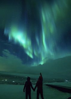 two people holding hands in front of an aurora bore over the ocean with bright green and blue lights