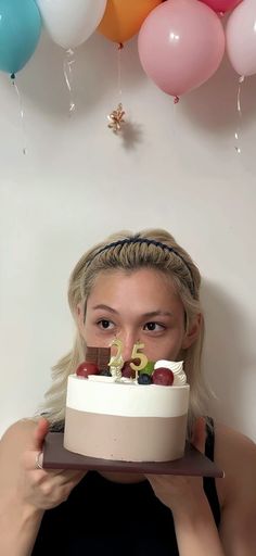 a woman holding a cake with candles in front of her face and balloons above her head