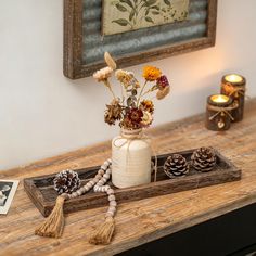 a vase with flowers and pine cones on a table in front of a framed picture