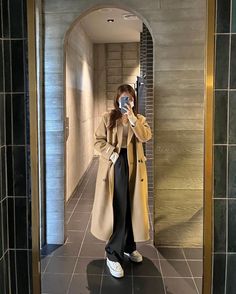 a woman taking a selfie in the mirror while wearing a trench coat and black pants