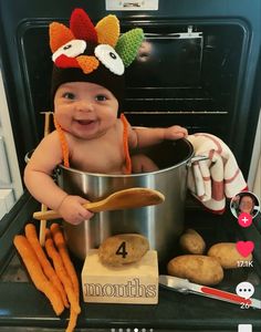 a baby wearing a turkey hat while sitting in an oven with carrots and potatoes