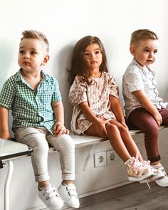 three children sitting on a white bench with their legs crossed and one child standing up