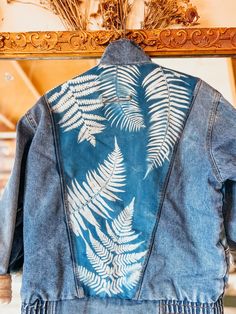 a blue jacket with white fern leaves on it hanging from a wooden framed frame in front of a mirror