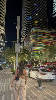 a woman is walking down the sidewalk in front of some tall buildings at night time