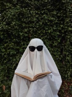 a white ghost with sunglasses and a book in front of a hedge covered in ivy