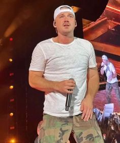 a male in a white shirt and camo pants is holding a microphone on stage