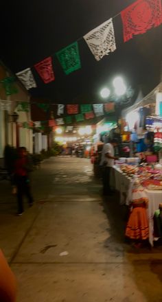 people are shopping at an outdoor market in the night time with colorful flags strung above them