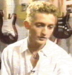 a man with blonde hair wearing a white shirt and tie sitting in front of guitars