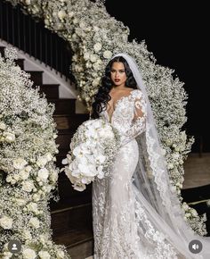 a woman in a wedding dress standing next to a flower covered archway with white flowers