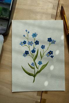 a watercolor painting of blue flowers on white paper next to a pen and pencil