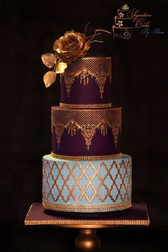 a three tiered cake with purple and blue icing, gold decorations and a rose on top