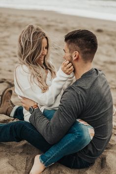 a man and woman sitting on top of a sandy beach next to the ocean holding each other