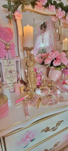 a dresser with pink flowers and candles on it, next to a mirror that is covered in gold trim