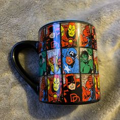 a black coffee mug with avengers characters on it sitting on a blanket in the sun