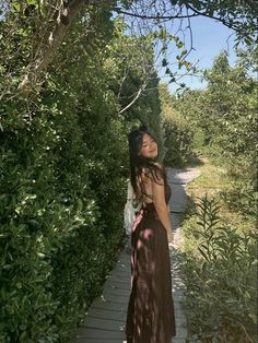 a woman in a long dress standing on a wooden walkway next to bushes and trees