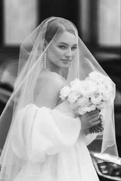 a woman in a wedding dress and veil holding a bouquet of flowers on the street