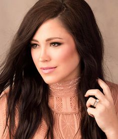 a woman with long dark hair wearing a pink top and holding her hand on her shoulder