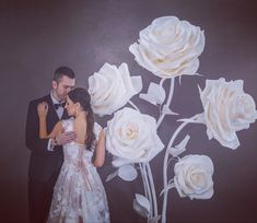 a couple standing next to each other in front of a wall with flowers on it