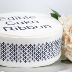 a white cake with blue and black designs on it, next to a flower that reads edible cake ribbon