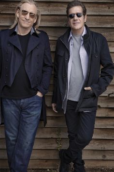 two men standing next to each other in front of a wooden wall with sunglasses on