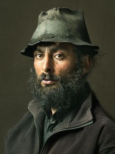 a man with a hat on his head and beard is looking at the camera while wearing a jacket