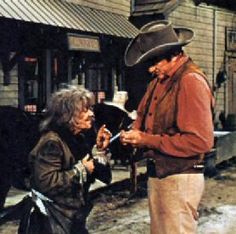 two men standing next to each other in front of a building and one is holding an umbrella