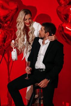 a man and woman sitting next to each other in front of red balloons holding wine glasses