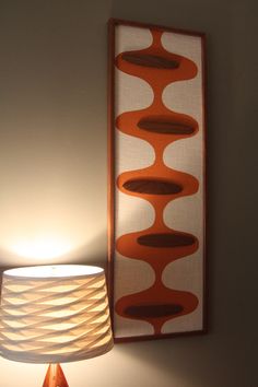 an orange lamp next to a wall hanging on the side of a white wall with a brown and red design