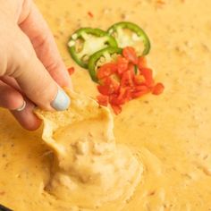 a hand dipping a tortilla chip into a bowl of dip with peppers and jalapenos on the side